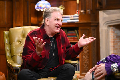 Michael Rapaport wearing a red jacket while sitting in a leather chair in The Traitors.