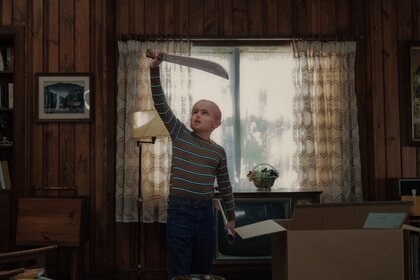 Callum Vinson as Jason Voorhees stands in a wood-paneled room holding a large machete overhead beside moving boxes and a television.