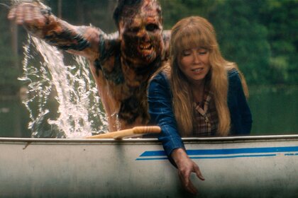Linda Cardellini as Pam Voorhees reaches over the side of a canoe as a burned figure rises from the water behind the canoe.
