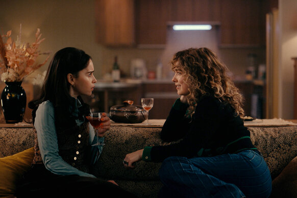 Bea (Emilia Clarke) and Twila (Haley Lu Richardson) sitting on a couch together during Ponies Season 1.