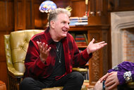 Michael Rapaport wearing a red jacket while sitting in a leather chair in The Traitors.