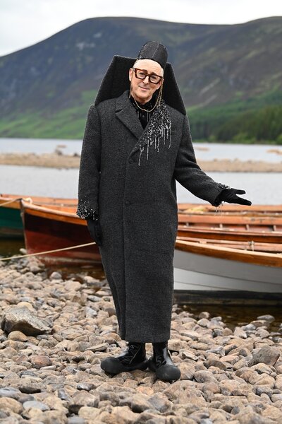 Alan Cumming stands near canoes on a rocky beach on The Traitors Episode 401.