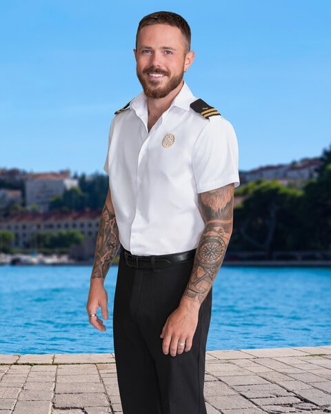 Nathan Gallagher wearing his yachting uniform while standing on a dock in front of the sea. and moutainside.