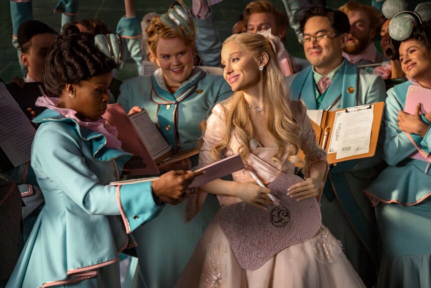 Glinda (Ariana Grande) smiles as students hold up their notebooks for her signature in Wicked For Good (2025).