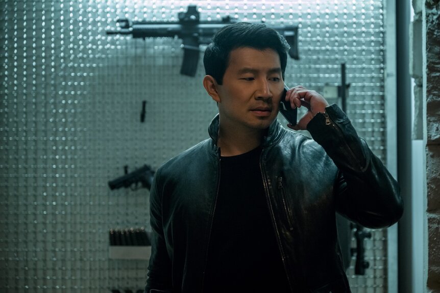 Alexander (Simu Liu) speaks on the phone in front of a gun wall on The Copenhagen Test Season 1.