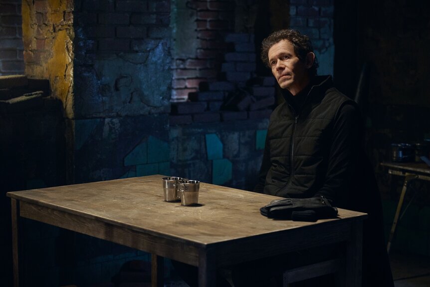Schiff (Adam Godley) sits at a wooden table with two metal cups and leather gloves on The Copenhagen Test Season 1.