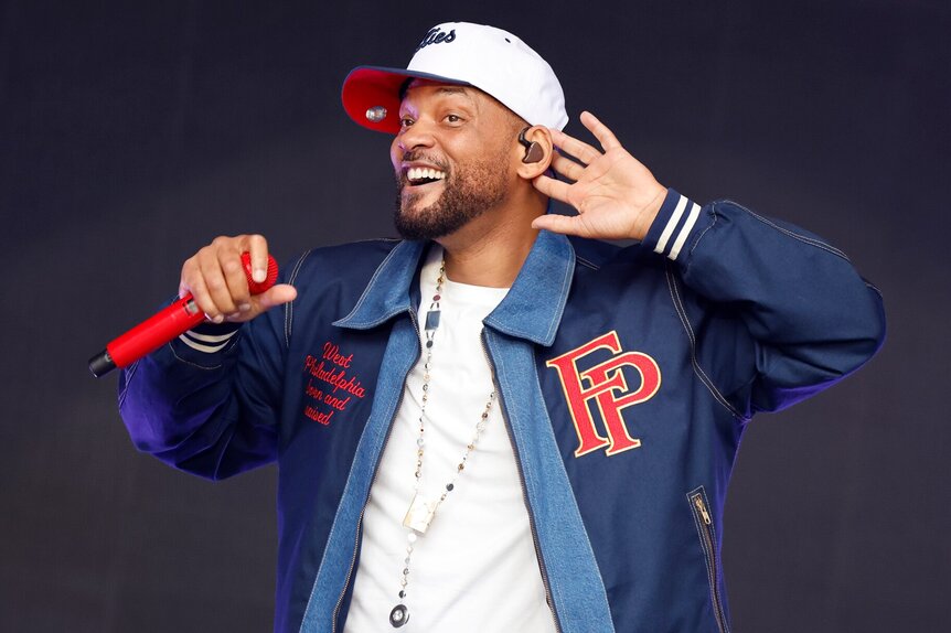 Will Smith holding a microphone while standing on stage wearing a baseball cap and a jacket