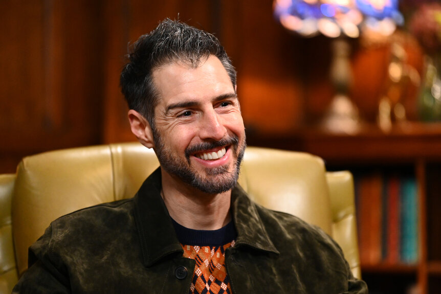 Rob Cesternino sitting in a library wearing a jacket and orange tee smiling during The Traitors