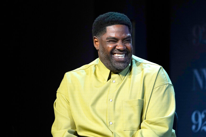 Ron Funches speaking on a stage wearing a yellow shirt