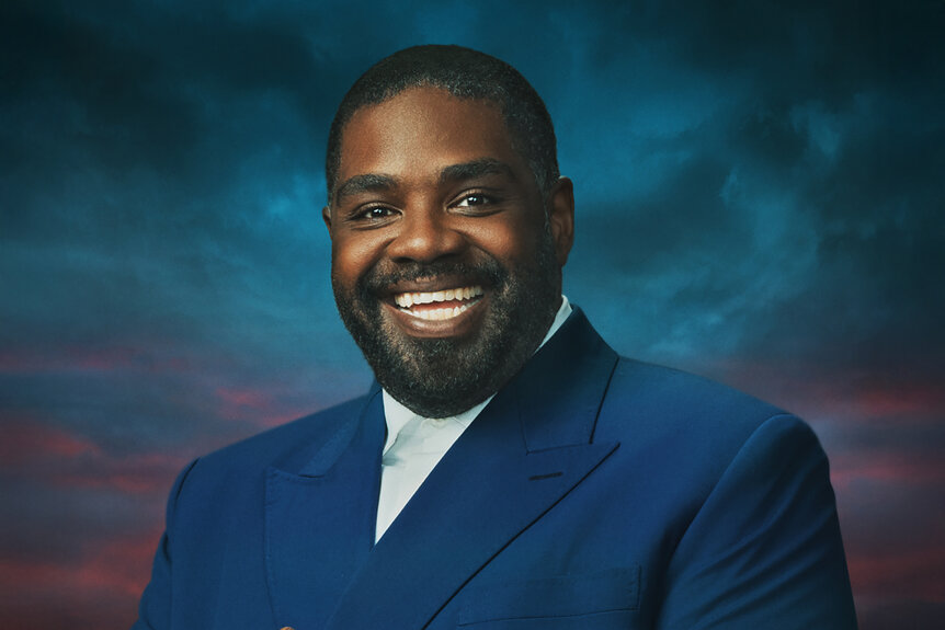 Ron Funches in a blue blazer and cloudy background for The Traitors Season 4.