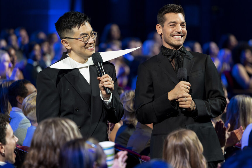 Bowen Yang and Matt Rogers smiling in the audience wearing black suits.