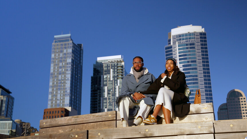 Mecca Amen and BelleJolie Force sitting on steps outside in front of large buildings.