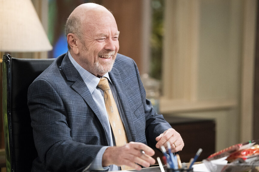 Phil Hendrie's character sitting behind a desk and laughing.