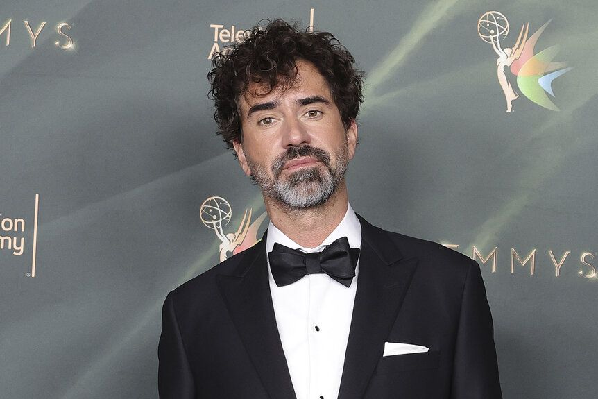 Hamish Linklater poses in a black tuxedo and bow tie in front of an Emmy Awards backdrop.
