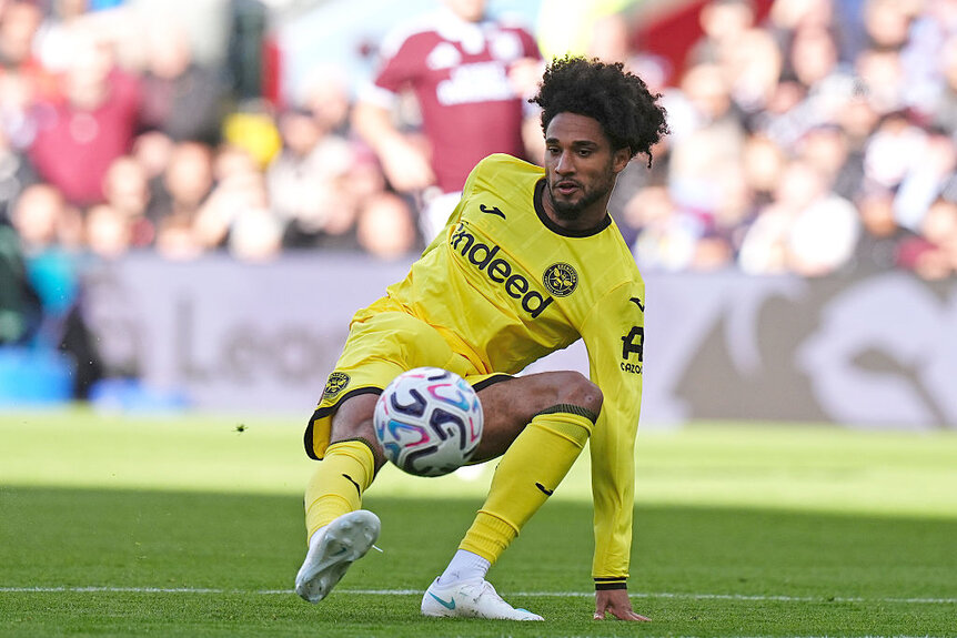Kevin Schade in a yellow Brentford kit slides on the pitch while attempting a shot during a Premier League match at Villa Park.