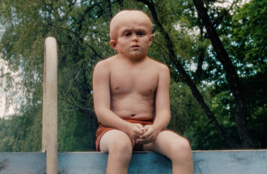 Callum Vinson as Jason Voorhees sits shirtless in red shorts on the edge of a dock with trees in the background.