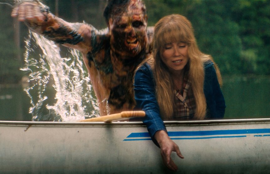 Linda Cardellini as Pam Voorhees reaches over the side of a canoe as a burned figure rises from the water behind the canoe.