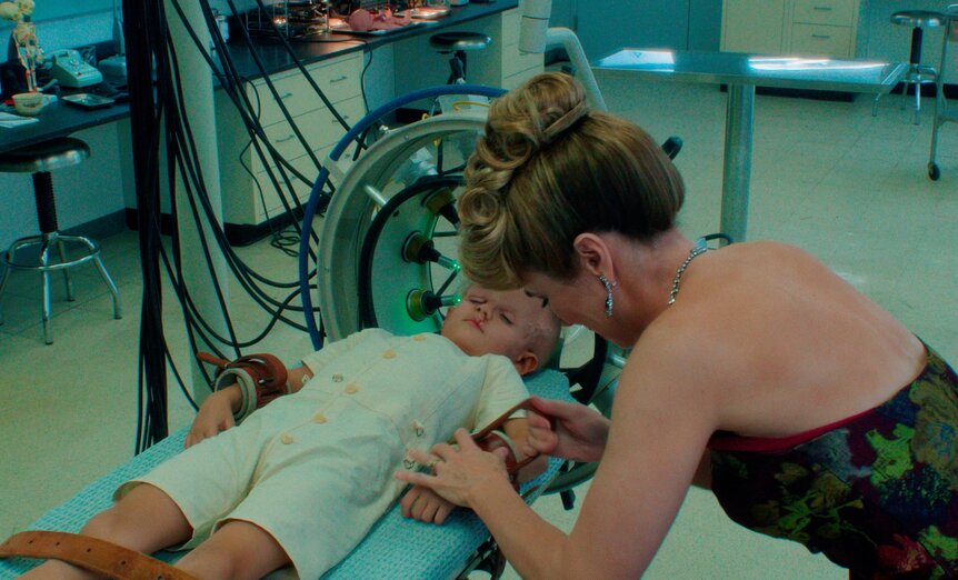 Linda Cardellini as Pam Voorhees leans over a child strapped to a medical table in a laboratory setting.